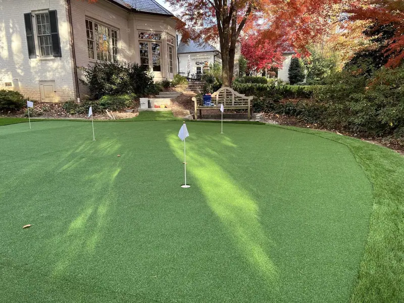 Large artificial turf putting green with a teak bench beside a stucco house