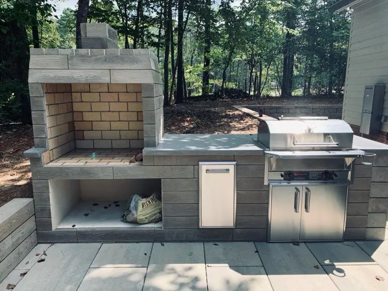 Outdoor kitchen with a built-in grill and fireplace on a paver patio in a wooded backyard